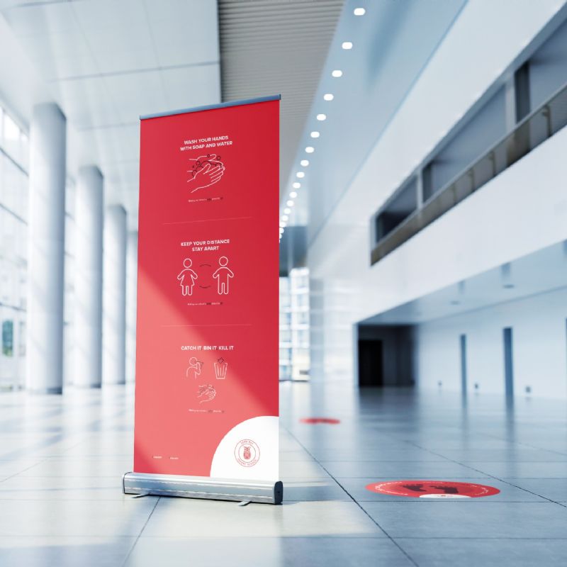 Red roller banner in an atrium with a red footprint floor sticker next to it. Banner depicts 3 COVID-19 awareness messages.